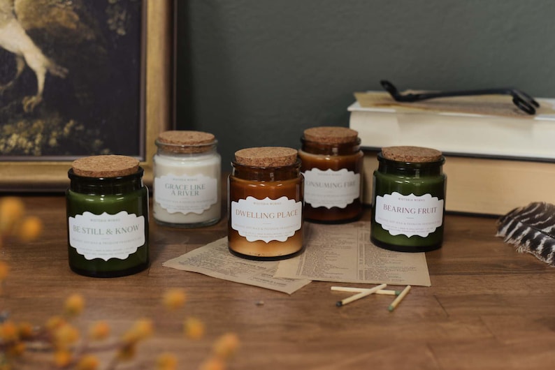 May include: Five glass jar candles with cork lids and labels. The candles are in shades of green, white, and brown. The labels read "Be Still & Know", "Grace Like a River", "Dwelling Place", "Final Mingling Fire", and "Bearing Fruit".