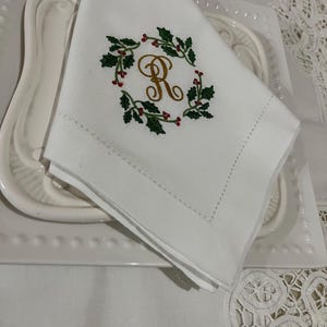 Peut inclure: Serviette en lin blanc avec une couronne de houx brodée et la lettre "R" en or. La serviette est pliée et posée sur une assiette blanche. La serviette a une bordure cousue.