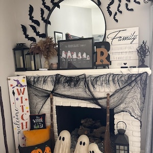 Może przedstawiać: Biały kominek z cegły ozdobiony na Halloween czarnymi nietoperzami, czarną siatką i tabliczką z napisem "Witamy Czarownica". Są też trzy białe duchy, miotła i poduszka z napisem "Czarownica jest w domu".