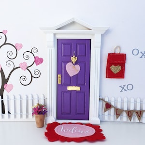 May include: A miniature purple door with a white frame and a pink heart wreath. The door has a gold doorknob and a small mailbox with a red door and a gold heart. The door is decorated with a welcome mat and a white picket fence. The background is a white wall with a black tree with pink hearts.