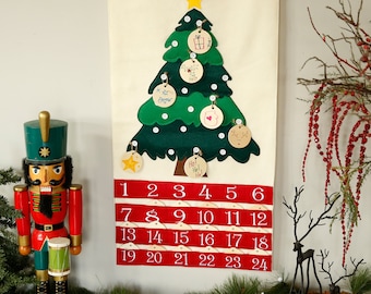 May include: A wall-hanging fabric advent calendar featuring a green felt Christmas tree with white buttons and hanging wooden ornaments, positioned above four rows of red pockets numbered one through twenty-four in white text.
