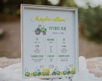 May include: A white framed printable poster with a green tractor illustration. The poster is decorated with yellow and green accents and includes details about a child's first year, including their height, weight, nickname, favorite foods, and books. The text on the poster reads "Brayden William, My First Year, August 11, 20XX".