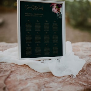May include: A dark green wedding seating chart with gold lettering. The chart is titled "Your Seat Awaits" and lists the names of guests for each table. The chart is decorated with a floral arrangement of burgundy and pink flowers.