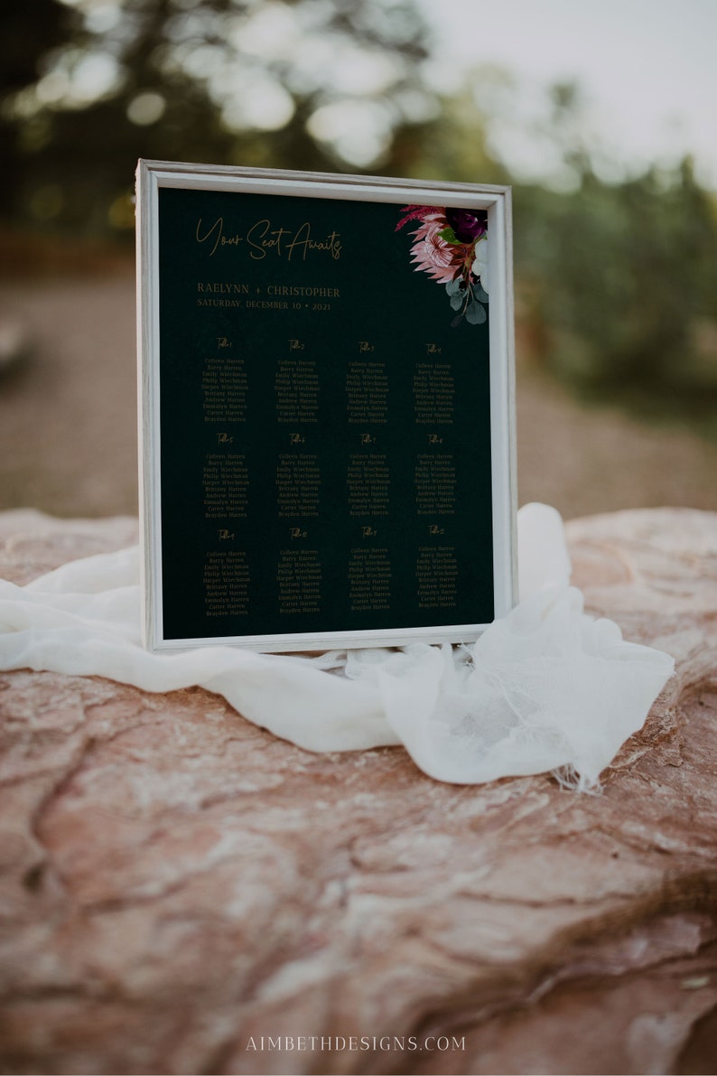 May include: A dark green wedding seating chart with gold lettering. The chart is titled "Your Seat Awaits" and lists the names of guests for each table. The chart is decorated with a floral arrangement of burgundy and pink flowers.