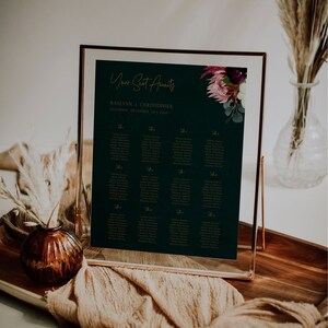 May include: A dark green wedding seating chart with gold lettering. The chart is framed in a gold frame and has a floral design in the top right corner. The text on the chart reads "Your Seat Awaits" and "Raelynn & Christopher, Saturday, December 10, 2023".