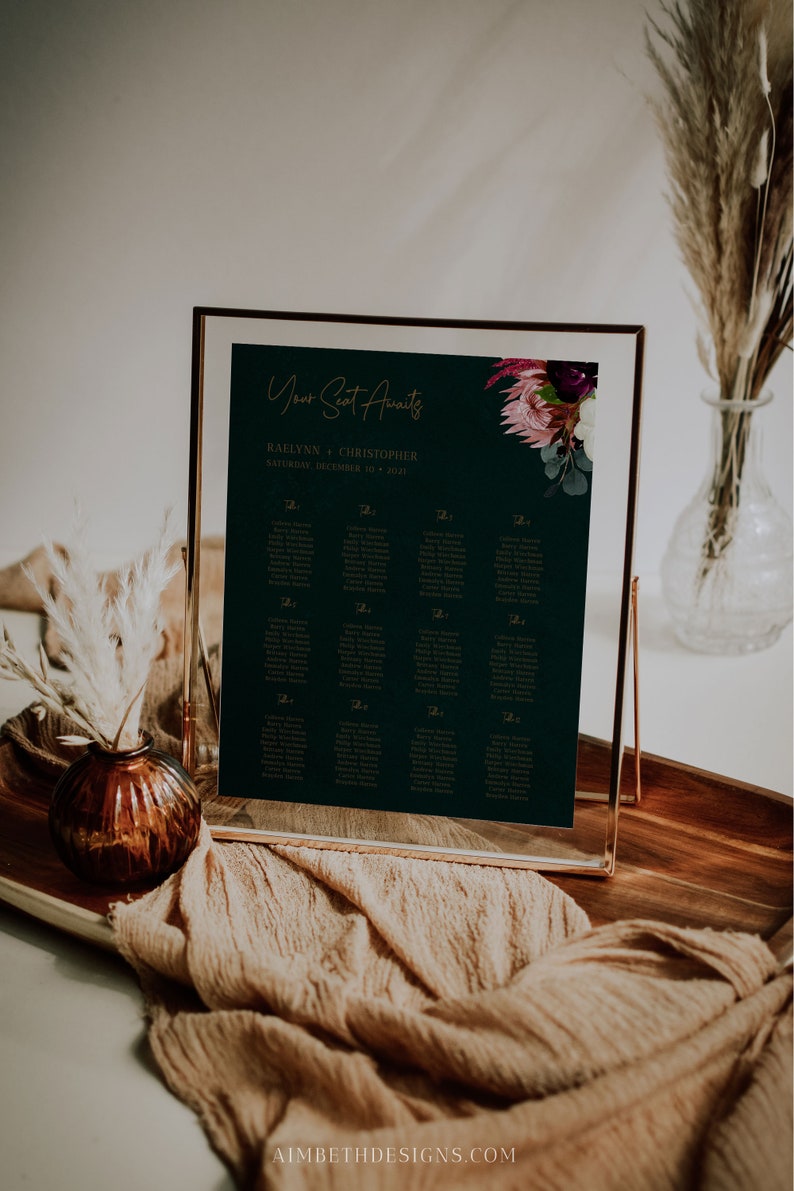 May include: A dark green wedding seating chart with gold lettering. The chart is framed in a gold frame and has a floral design in the top right corner. The text on the chart reads "Your Seat Awaits" and "Raelynn & Christopher, Saturday, December 10, 2023".