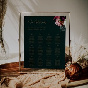 May include: A dark green wedding seating chart with gold lettering. The chart is framed in a gold frame and features a floral design with pink and purple flowers. The text on the chart reads "Your Seat Awaits" and "Raelynn + Christopher, Saturday, December 10, 2021".