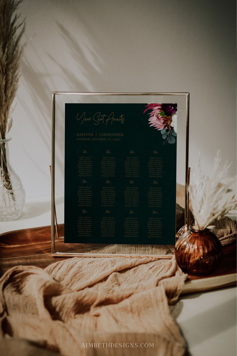 May include: A dark green wedding seating chart with gold lettering. The chart is framed in a gold frame and features a floral design with pink and purple flowers. The text on the chart reads "Your Seat Awaits" and "Raelynn + Christopher, Saturday, December 10, 2021".