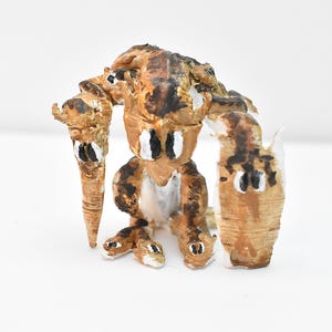 May include: A small, brown, ceramic sculpture of a creature with multiple limbs and large, black eyes. The creature is sitting on a white surface.