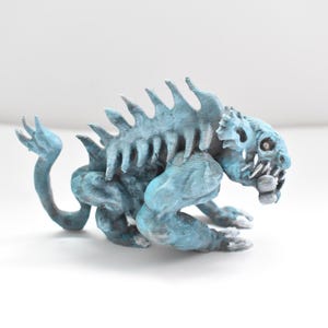 May include: A light blue, stylized creature figurine with a spiky spine, sharp teeth, and a curled tail. The figurine is posed in a crouching position, with its front paws extended. The creature has a textured surface and is set against a white background.