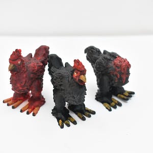 May include: Three toy chickens, each with a different color scheme. One is red, one is black, and one is black with red accents. The chickens are all standing on a white surface.