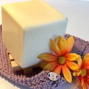 Peut inclure: Une grande barre de savon blanche carrée repose sur un porte-savon en bois. Le porte-savon est sur une surface en tricot violet. Deux fleurs orange et jaunes sont au premier plan.