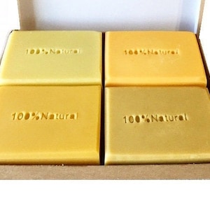 May include: Four rectangular bars of soap in shades of yellow are presented in a cardboard box. Each bar is stamped with the words "100% Natural". The soap bars are arranged in a neat, organised manner inside the box.
