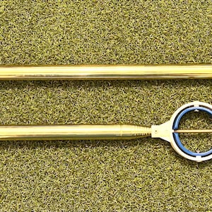 May include: Two gold-colored golf putting training aids with blue handles. The top of each aid has a white ring with a blue center and a gold rod.