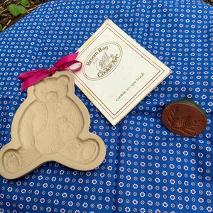 May include: A vintage Brown Bag Cookie Art cookie cutter in the shape of a bear holding a smaller bear. The cutter is made of a light brown ceramic material and is sitting on a blue and white patterned fabric. The cutter is accompanied by a vintage Brown Bag Cookie Art recipe book and a brown ceramic cookie stamp with a pine cone design.