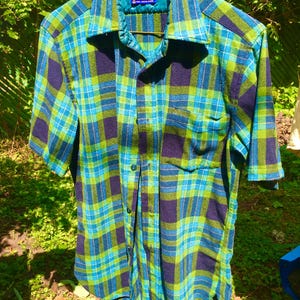 May include: A short-sleeved plaid shirt in shades of blue, green, and navy. The shirt has a collared neckline and a single chest pocket. The fabric appears to be a woven material. The shirt is hanging outdoors.