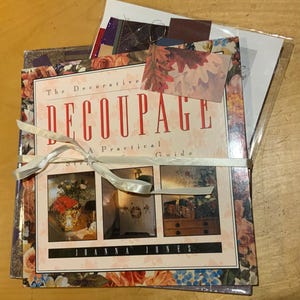 May include: A stack of books and ephemera tied with a white ribbon. The book cover reads "The Decorative Decoupage: A Practical Guide" by Joanna Jones. Floral and botanical designs adorn the book covers and a decorative tag with fall leaves.