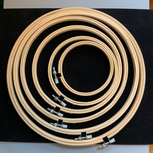 Op de afbeelding: Een verzameling houten borduurringen van verschillende formaten, in elkaar genest. Elke ring is gemaakt van lichtgekleurd hout en heeft een metalen spanmechanisme. De ringen worden getoond op een donkere ondergrond.