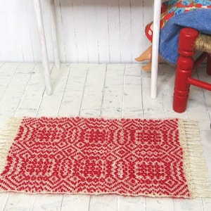 May include: A small, rectangular red and cream-colored woven rug with a geometric pattern. The rug has a fringed edge and is placed on a white wooden floor. A doll sits on a red chair near a small table.