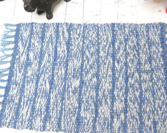 May include: A rectangular woven rug featuring a vertical striped pattern in shades of blue and white. The rug has a textured surface with fringe detailing along the two shorter edges, creating a consistent and repeating linear design throughout.