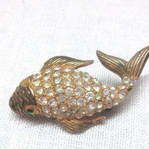 May include: A gold-toned metal brooch shaped like a fish, featuring a body covered in clear, round rhinestones. The head, fins, and tail are textured with etched lines, and the eye is accented with a small, singular green stone.