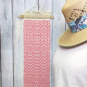 May include: A pink and white woven wall hanging with a geometric pattern, displayed on a wooden loom. The textile has a fringed bottom edge and is suspended from a decorative hanger. The background is a weathered, light-colored wooden wall.