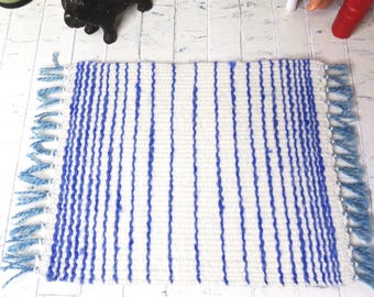 May include: A miniature rug with a white background and vertical blue stripes. The rug has fringed edges and is placed on a white surface. A black dog figurine and a decorative vase are also visible.
