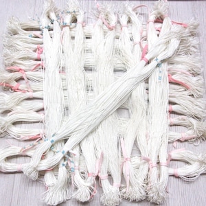 May include: A collection of white thread bundles, tied with pink and blue string, arranged in a woven pattern. The threads are tightly wound, creating a textured appearance. The background is a light-colored wooden surface.
