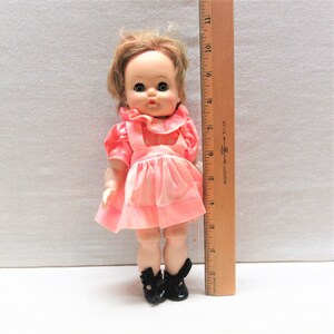 May include: A vintage doll with blonde hair, wearing a pink dress and black shoes. The doll has a sweet expression and is approximately 10 inches tall, as measured by a wooden ruler.
