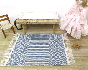 May include: A miniature blue and white patterned rug with fringe. The rug features a geometric design with a striped center and decorative edges. A small wooden table and chair are in the background, along with a doll.