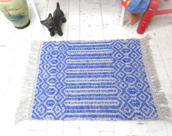 May include: A small, rectangular blue and white woven rug with a geometric pattern. The rug has a fringe on the short ends. The rug is likely for a dollhouse or miniature setting.