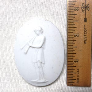May include: White oval-shaped ceramic plaque with a bas-relief of a figure playing a flute. The figure is wearing a short tunic. A wooden ruler with inch markings is next to the plaque.