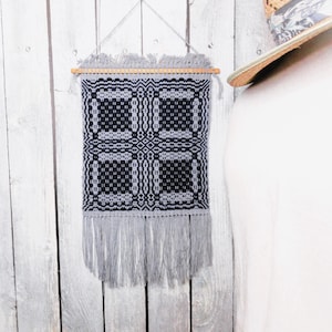 May include: A black and white woven wall hanging with a geometric pattern and gray fringe. The hanging is mounted on a wooden dowel rod.
