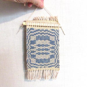 May include: A small woven wall hanging with a blue and white geometric pattern. The hanging has a wooden dowel at the top and a fringe at the bottom.