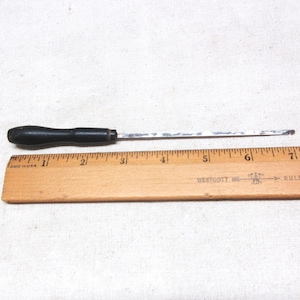 May include: A vintage metal tool with a black wooden handle, approximately 7 inches long, lies next to a wooden ruler. The ruler is marked with inches and the text "WESTCOTT RULER". The tool appears to be a type of awl or similar instrument.