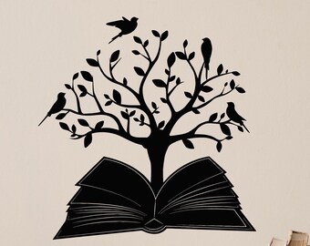 May include: A black silhouette of a tree with leaves and birds growing out of an open book. The image is a wall decal. The background is a light beige color. The decal is on a wall above a dark brown wooden table with books and decorative items.