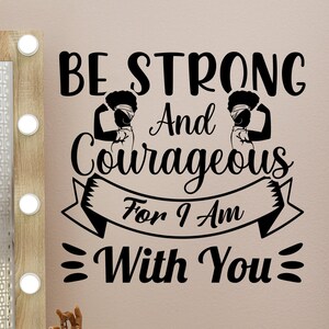 Puede incluir: Un espejo de tocador con marco de madera con bombillas, un maniquí de mano de madera y una calcomanía de pared. La calcomanía negra dice "BE STRONG And Courageous For I Am With You". El fondo es de color rosa claro.