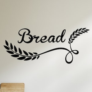 May include: Black and white vinyl wall decal with the word "Bread" and wheat stalks. The decal is on a white wall above a white kitchen cabinet with gold handles.