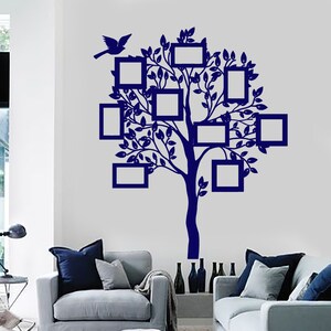 Op de afbeelding: Een blauwe silhouet van een boom met takken en bladeren, met acht vierkante frames die aan de takken hangen. Een vogel zit op een tak. De boom bevindt zich op een witte muur in een woonkamer met twee grijze banken.