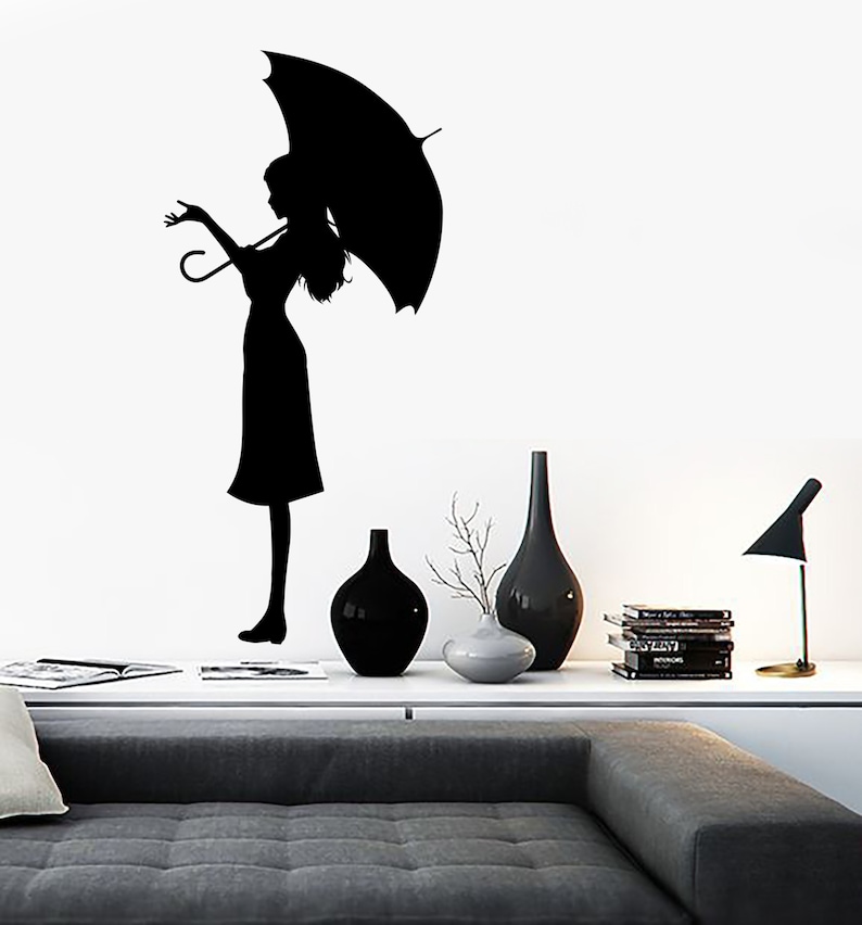 May include: Black silhouette wall decal of a person holding an umbrella. The decal is on a white wall with a couch and decorative items in the foreground.