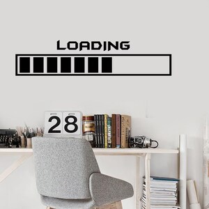 May include: A black and white loading bar graphic with the word "LOADING" above it. The graphic is on a white wall. A desk with a chair, books, and a calendar is in the background.