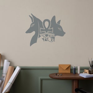 Op de afbeelding: Een grijze muursticker met twee gestileerde dierenkoppen die een ankh-symbool met het oog van Horus flankeren. De ankh is een symbool van het leven en het oog van Horus staat voor bescherming en gezondheid.
