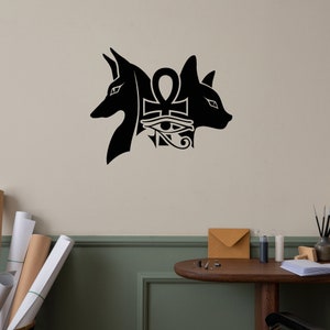 Op de afbeelding: Zwarte en witte muursticker met twee gestileerde dierenkoppen die een ankh-symbool met het oog van Horus flankeren.