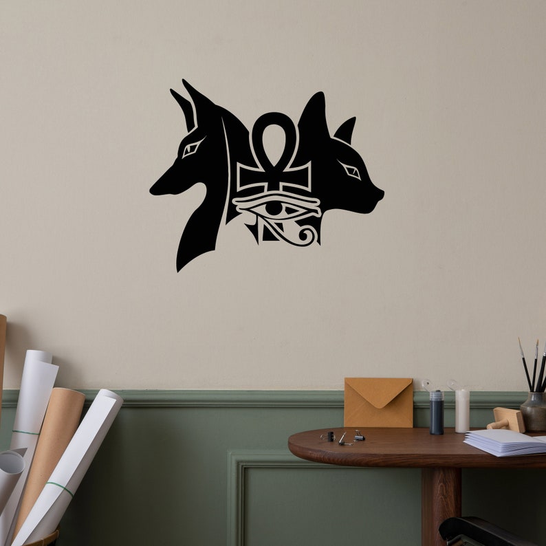 Op de afbeelding: Zwarte en witte muursticker met twee gestileerde dierenkoppen die een ankh-symbool met het oog van Horus flankeren.