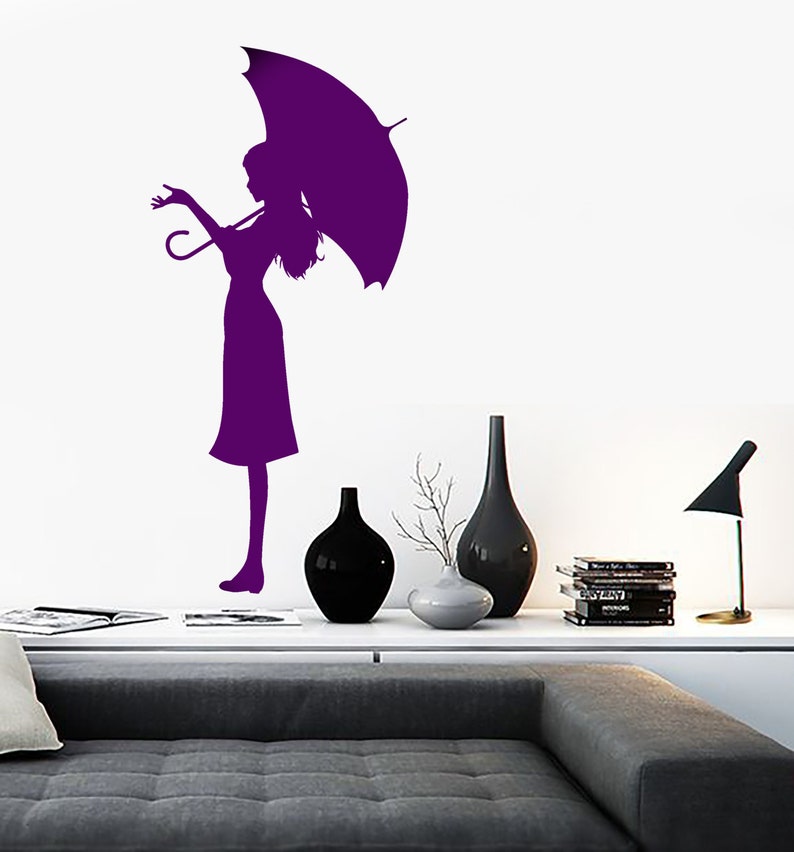 May include: Purple silhouette of a person holding an umbrella, standing on a white wall. The person is wearing a dress and has long hair. The image is a wall decal.