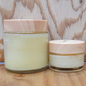 Könnte beinhalten: Zwei gläserne Behälter mit Holzdeckeln, gefüllt mit einer hellgelben Creme. Der größere Behälter befindet sich links und der kleinere rechts.
