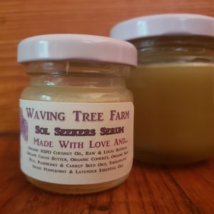 May include: Two small glass jars with white lids. The jar on the left contains a light yellow serum and has a label that reads "Waving Tree Farm Sol Seekers Serum Made With Love And... Organic RSPO Coconut Oil, Raw & Local Beeswax, Organic Cocoa Butter, Organic Comfrey, Organic Sea Salt, Raspberry & Carrot Seed Oils, Therapeutic Grade Peppermint & Lavender Essential Oils."