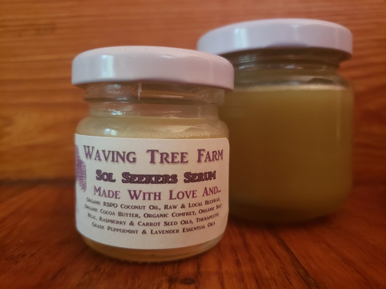 May include: Two small glass jars with white lids. The jar on the left contains a light yellow serum and has a label that reads "Waving Tree Farm Sol Seekers Serum Made With Love And... Organic RSPO Coconut Oil, Raw & Local Beeswax, Organic Cocoa Butter, Organic Comfrey, Organic Sea Salt, Raspberry & Carrot Seed Oils, Therapeutic Grade Peppermint & Lavender Essential Oils."