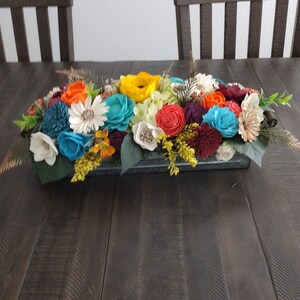 May include: A colorful floral arrangement in a rectangular galvanized metal container. The flowers are a mix of bright colors, including orange, yellow, blue, and white. The arrangement sits on a dark wooden table, with two wooden chairs in the background.