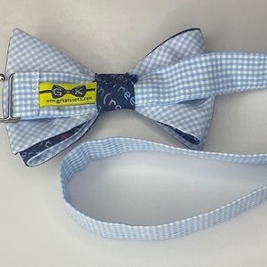 May include: A blue and white gingham bow tie with a blue and white gingham strap. The bow tie has a yellow label with the text "GK" and "www.greatknots.com".